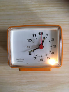 Réveil Vedette rétro en plastique orange, aiguilles fluo, bouton poussoir pour éteindre la sonnerie, pile 1,5V non incluse, fonctionne parfaitement, dimensions :  L 10, H 9, P 5,5 cm.