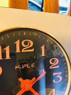Horloge murale Kiple, support carré en bois teint en gris irisé, chiffres et aiguilles orange pop, mécanisme quartz.