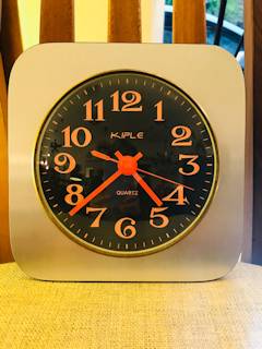 Horloge murale Kiple, support carré en bois teint en gris irisé, chiffres et aiguilles orange pop, mécanisme quartz.